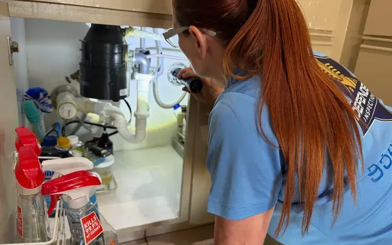 Inspector using a flashlight to check supply lines and the trap under a kitchen sink